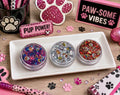 Three jars of Paw Patrol-themed hair glitter in pink, silver, and red-gold with paw print confetti